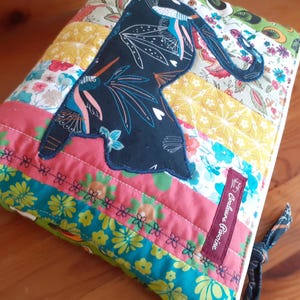 May include: A quilted fabric pouch with a dark blue bird applique. The pouch features a patchwork design with floral and geometric patterns in shades of pink, yellow, and teal. A small burgundy label with text is sewn on the side.