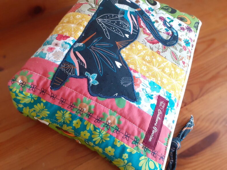 May include: A quilted fabric pouch with a dark blue bird applique. The pouch features a patchwork design with floral and geometric patterns in shades of pink, yellow, and teal. A small burgundy label with text is sewn on the side.