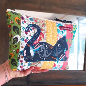 May include: A patchwork cosmetic bag with a navy blue cat applique. The bag features a variety of colorful floral and patterned fabrics, including green, yellow, and pink. A black tassel is attached to the zipper.