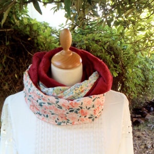 Peut inclure: Écharpe infinie bordeaux avec un motif floral rose, vert et jaune. L'écharpe est superposée à un haut blanc et une tête de mannequin en bois. L'arrière-plan présente un feuillage vert.