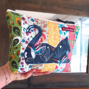 Peut inclure: Une trousse de maquillage patchwork avec un appliqué de chat bleu marine. Le sac présente une variété de tissus floraux et à motifs colorés, dont du vert, du jaune et du rose. Une pompon noir est attaché à la fermeture éclair.