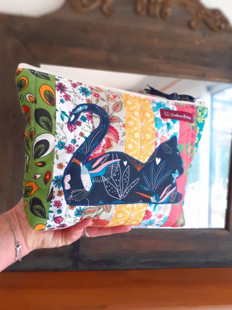 May include: A patchwork cosmetic bag with a navy blue cat applique. The bag features a variety of colorful floral and patterned fabrics, including green, yellow, and pink. A black tassel is attached to the zipper.