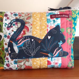 May include: A quilted fabric pouch with a patchwork design. The pouch features a dark blue cat silhouette with floral and hummingbird details. The background is a mix of floral and solid fabric panels in various colors.