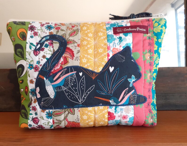 May include: A quilted fabric pouch with a patchwork design. The pouch features a dark blue cat silhouette with floral and hummingbird details. The background is a mix of floral and solid fabric panels in various colors.