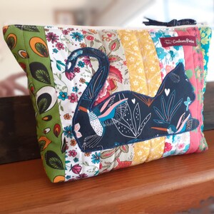 May include: A quilted pouch with a patchwork design in various floral patterns and colors. A dark blue cat silhouette is appliqued on the front, decorated with white floral details and small birds. The pouch has a zipper closure.