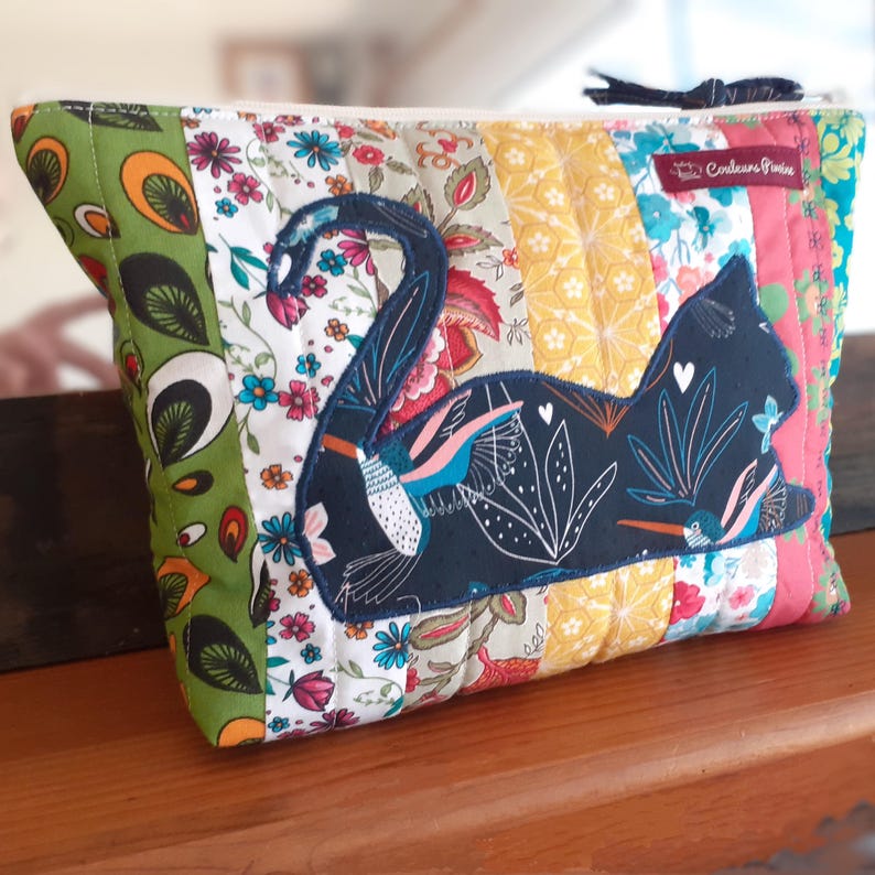 May include: A quilted pouch with a patchwork design in various floral patterns and colors. A dark blue cat silhouette is appliqued on the front, decorated with white floral details and small birds. The pouch has a zipper closure.