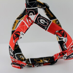 Pu&ograve; includere: Un'imbracatura per cani rossa, nera e bianca con un motivo ripetuto del logo di un bulldog. L'imbracatura ha una fibbia nera e cinghie regolabili. Lo sfondo &egrave; bianco.