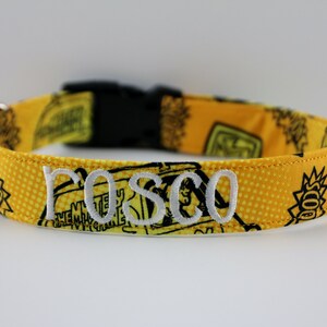 May include: Yellow dog collar with black graphics and the embroidered name "ROSCO" in white. The collar has a black plastic buckle and a metal D-ring for leash attachment. The fabric features a repeating pattern of cartoon characters.