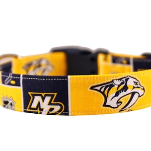 Puede incluir: Un collar de perro amarillo y azul con el logotipo de los Nashville Predators. El collar tiene una hebilla negra.