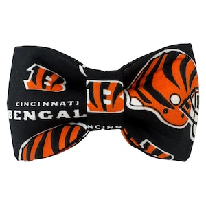 Könnte beinhalten: Schwarze Fliege mit orange-weißem Design der Cincinnati Bengals. Die Fliege zeigt das Logo, den Helm und den Text "CINCINNATI BENGALS". Dieses Accessoire eignet sich für besondere Anlässe oder zur Unterstützung des Teams.