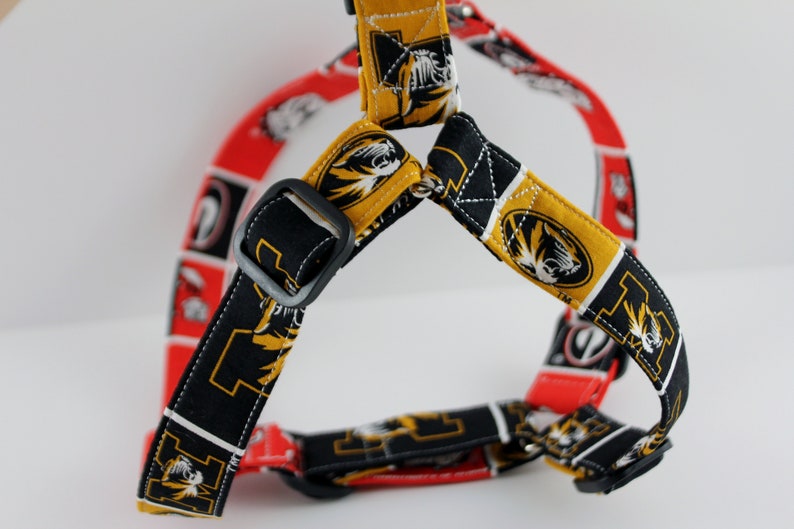 Pu&ograve; includere: Un'imbracatura per cani con un motivo ripetuto in nero, oro e rosso. L'imbracatura ha cinghie regolabili e una fibbia nera. Il design del tessuto include un logo di tigre e altre grafiche.