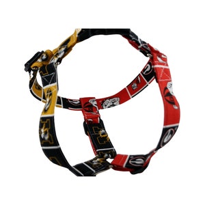 Pu&ograve; includere: Un'imbracatura per cani con un motivo nero, rosso e oro. L'imbracatura presenta un motivo ripetuto di un logo bulldog e le lettere "G" e "A". L'imbracatura ha cinghie regolabili e una fibbia in plastica nera.