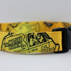 May include: Yellow dog collar featuring a black and white cartoon design with the text "THE MYSTERY MACHINE". The collar has a black plastic buckle and a matching adjustable strap.