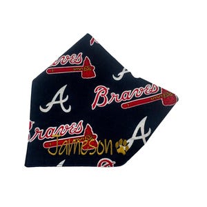 Op de afbeelding: Een marineblauwe bandana met een witte pootprint en de tekst "Jameson" in goud. De bandana is versierd met een herhalend patroon van het Atlanta Braves-logo, met een tomahawk en de woorden "Braves" en "A".