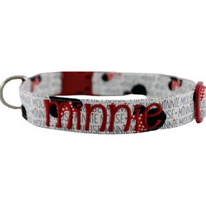 May include: A white dog collar with the name "Minnie" embroidered in red and black. The collar features a pattern of Minnie Mouse silhouettes and text. It has a metal loop and a red buckle.