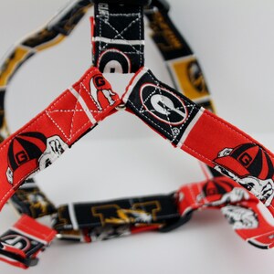 Pu&ograve; includere: Un'imbracatura per cani con un motivo rosso e nero, con una grafica di bulldog. L'imbracatura ha cinghie regolabili ed &egrave; realizzata in tessuto. Il design include la lettera "G" e altre grafiche a tema.