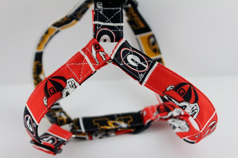 Pu&ograve; includere: Un'imbracatura per cani con un motivo rosso e nero, con una grafica di bulldog. L'imbracatura ha cinghie regolabili ed &egrave; realizzata in tessuto. Il design include la lettera "G" e altre grafiche a tema.