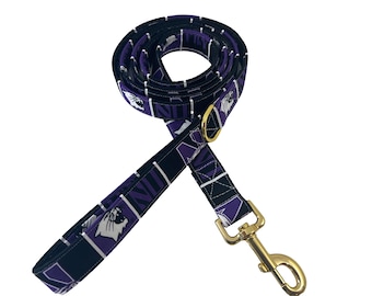 May include: A rolled-up dog leash with a repeating pattern of a purple and white wildcat logo and the word "WILDCATS" on a navy blue background. The leash has a gold-colored metal clasp and ring.