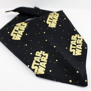 May include: Black bandana with gold "STAR WARS" text and small gold dots. The bandana is folded in a triangle shape. The Star Wars logo is repeated multiple times on the fabric.