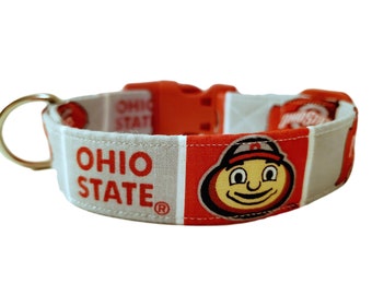 May include: A white and red dog collar with the words "OHIO STATE" in red. The collar has an orange buckle and a gold-coloured metal ring. The Ohio State Buckeyes mascot is also featured on the collar.