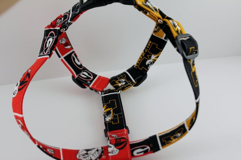 Pu&ograve; includere: Un'imbracatura per cani con un motivo rosso, nero e bianco con il logo dei Bulldogs dell'Universit&agrave; della Georgia e un motivo nero, oro e bianco con il logo dei Tigers del Missouri. Ha fibbie in plastica nera regolabili e un manico nero.