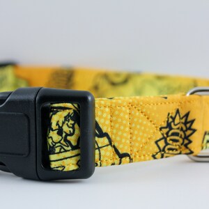 May include: Yellow dog collar with a black plastic buckle and a silver metal D-ring. The collar features a repeating cartoon graphic pattern in black and yellow. The collar is made of fabric.