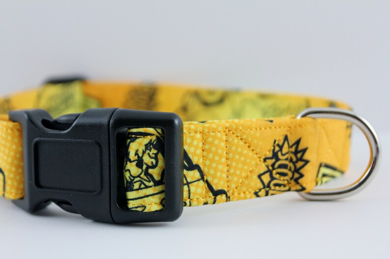 May include: Yellow dog collar with a black plastic buckle and a silver metal D-ring. The collar features a repeating cartoon graphic pattern in black and yellow. The collar is made of fabric.
