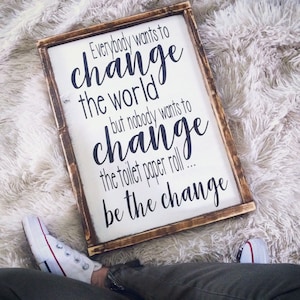 May include: A rectangular wooden sign with the text "Everybody wants to change the world but nobody wants to change the toilet paper roll... be the change" in black script. The sign has a white background and a brown frame.