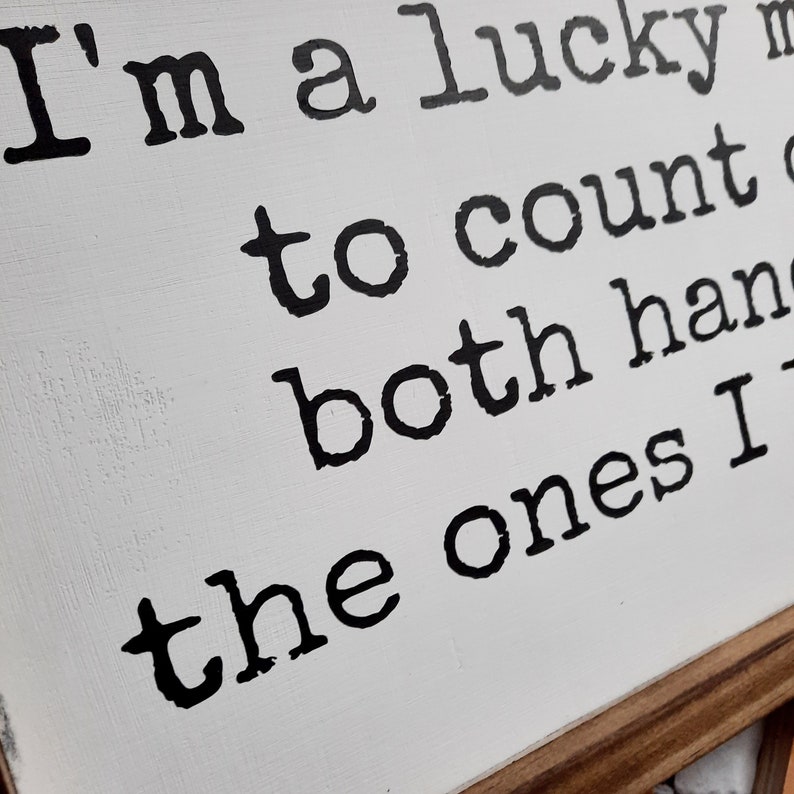 May include: White wooden sign with black text that reads "I'm a lucky man to count both hands the ones I"