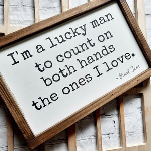 May include: A wooden sign with a white background and brown frame. The sign features a quote from Pearl Jam: "I'm a lucky man to count on both hands the ones I love."