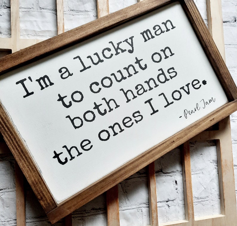May include: A wooden sign with a white background and brown frame. The sign features a quote from Pearl Jam: "I'm a lucky man to count on both hands the ones I love."
