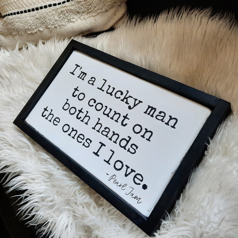 May include: Black framed sign with white background and black text that reads "I'm a lucky man to count on both hands the ones I love. - Pearl Jam"