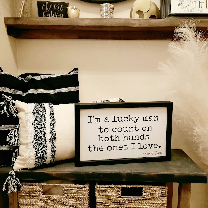 May include: Black framed wooden sign with white background and black text that reads "I'm a lucky man to count on both hands the ones I love. -Pearl Jam".