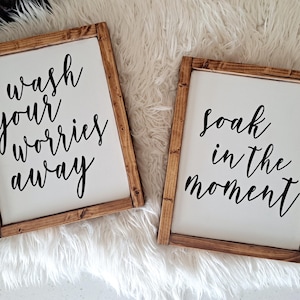 May include: Two framed wooden signs with white backgrounds and black cursive text. One sign reads "wash your worries away," and the other says "soak in the moment." The frames are a warm brown color.