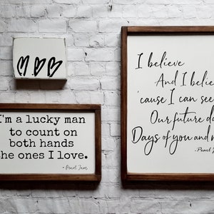 May include: Three wooden signs with white backgrounds and black text. The top sign has three hearts drawn on it. The middle sign says "I'm a lucky man to count on both hands the ones I love. -Pearl Jam". The bottom sign says "I believe And I believe 'cause I can see Our future days Days of you and me -Pearl Jam".