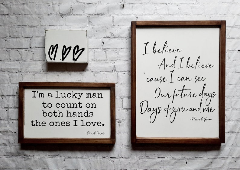 May include: Three wooden signs with white backgrounds and black text. The top sign has three hearts drawn on it. The middle sign says "I'm a lucky man to count on both hands the ones I love. -Pearl Jam". The bottom sign says "I believe And I believe 'cause I can see Our future days Days of you and me -Pearl Jam".