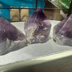 May include: Three amethyst crystal points, each with a unique shape and purple hue, are displayed on a white surface. The crystals are polished and have a natural, earthy look.