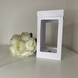 May include: A white rectangular gift box with a large rectangular window. The box is next to a bouquet of white peonies. The box is made of cardboard and has a scalloped edge on the top.