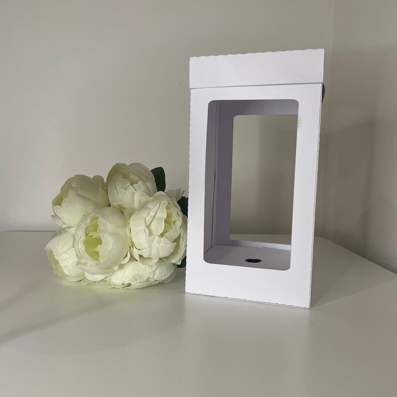 May include: A white rectangular gift box with a large rectangular window. The box is next to a bouquet of white peonies. The box is made of cardboard and has a scalloped edge on the top.