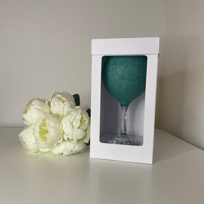 May include: A teal glitter-covered wine glass in a white box. The glass has a clear stem and base. A bouquet of white flowers is to the left of the box. The background is a plain white wall and surface.