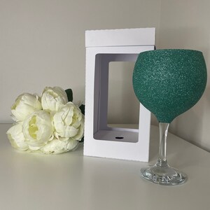 May include: A green glitter-covered wine glass with a clear stem and base. A white box with a window is behind the glass. A bouquet of white flowers is to the left of the box and glass.