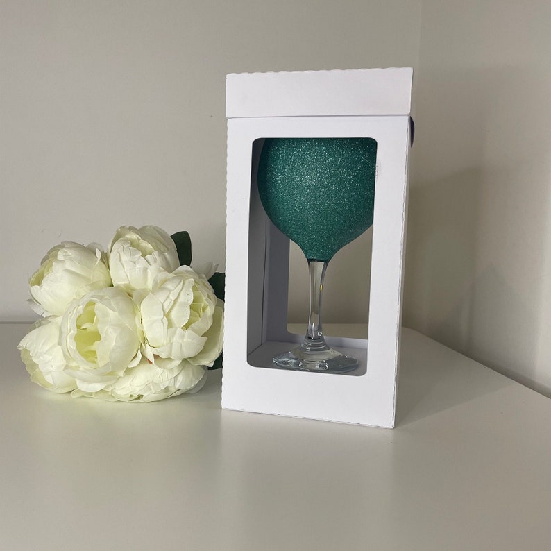 May include: A teal glitter-covered wine glass is displayed in a white box. The glass has a long stem and a round bowl. A bouquet of white peonies is to the left of the box. The background is a plain white wall and a white surface.