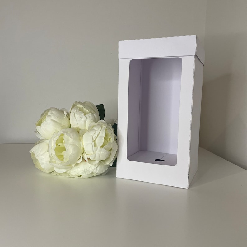 May include: A white rectangular gift box with a large rectangular window. A bouquet of white peonies with green stems and leaves is to the left of the box. The box is on a white surface.
