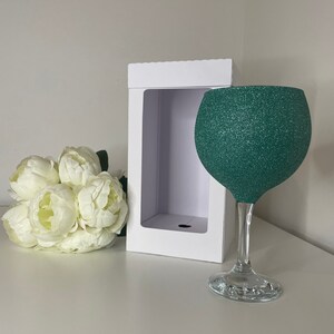 May include: A large, clear-stemmed wine glass with a teal glitter-covered bowl. A white gift box with a window is behind the glass. A bouquet of white flowers sits to the left of the glass. The glass is on a white surface.