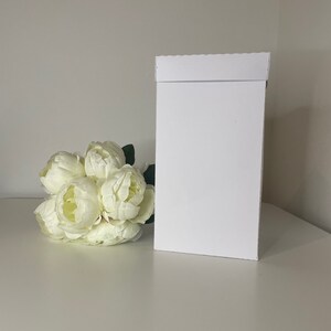 May include: A white rectangular gift box with a folded top, next to a bouquet of white peonies. The box is plain and stands upright on a white surface. The flowers are clustered together with green leaves.