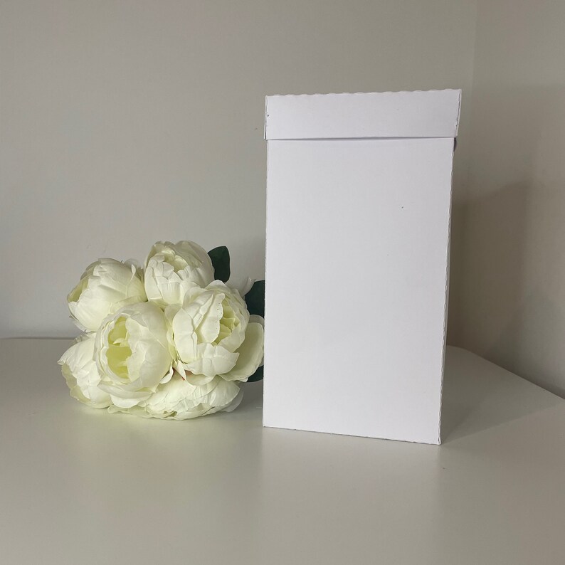 May include: A white rectangular gift box with a folded top, next to a bouquet of white peonies. The box is plain and stands upright on a white surface. The flowers are clustered together with green leaves.