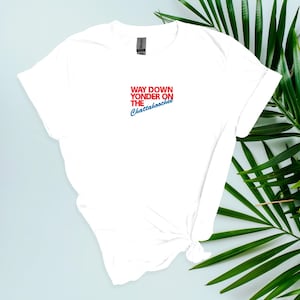 Puede incluir: Camiseta blanca con el texto "WAY DOWN YONDER ON THE Chattahoochee" en rojo y azul. La camiseta está anudada en la parte inferior y tiene mangas cortas. Hojas verdes en el fondo.