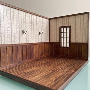 Könnte beinhalten: Ein Miniatur-Raumdiorama mit Holzboden und Wänden. Die Wände sind mit einer gemusterten Tapete und dunklen Holzvertäfelungen versehen. Zwei Wandleuchten sind an der Wand angebracht. Ein kleines Fenster ist an der Rückwand zu sehen. Der Raum ist mit einem dunklen Holzrahmen konstruiert.