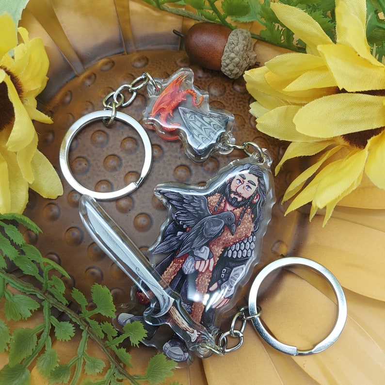 Może przedstawiać: Srebrny brelok do kluczy z czarno-białym obrazkiem mężczyzny z krukiem na ramieniu. Mężczyzna ma na sobie brązową sk&oacute;rzaną kurtkę i brodę. Brelok zawiera r&oacute;wnież wisiorkę w kształcie czerwono-pomarańczowego smoka.