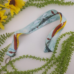 May include: A light blue lanyard with a repeating pattern of a mountain range and red and yellow dragons. The lanyard has a silver-colored metal clasp. The background includes yellow sunflowers and green foliage.